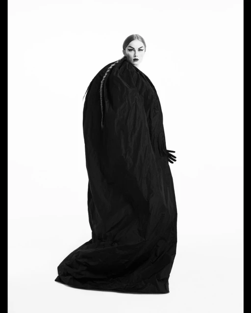 Woman wearing an oversized, draped black garment in a fashion-style shot against a stark white background. Dramatic styling and makeup.