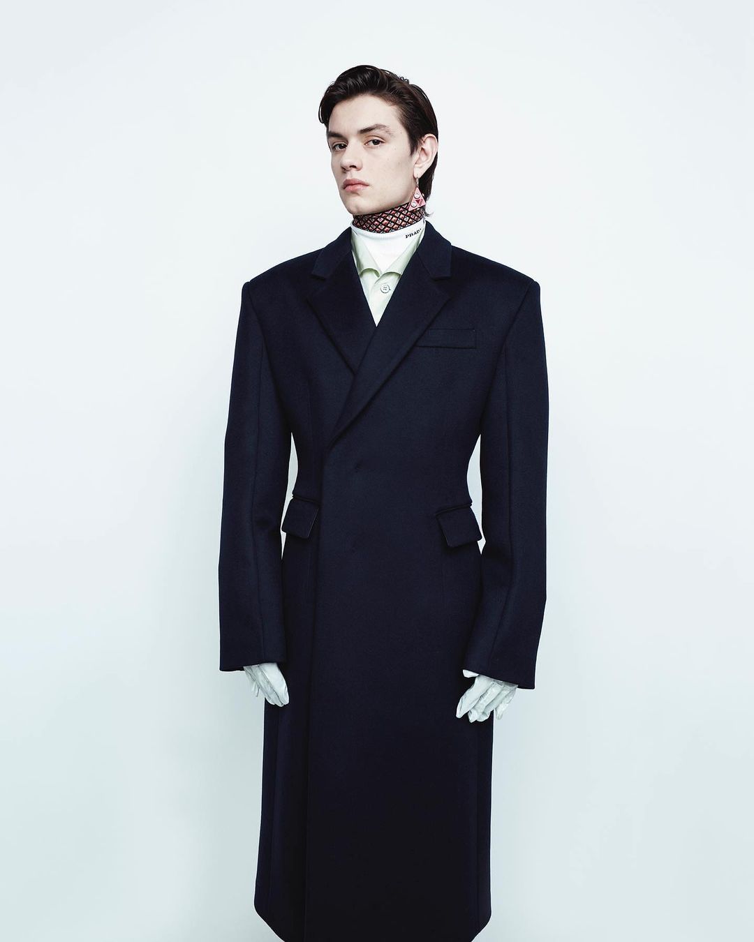 Fashion portrait of a young adult male model in an oversized navy coat, white gloves, and patterned neck scarf against a white backdrop.