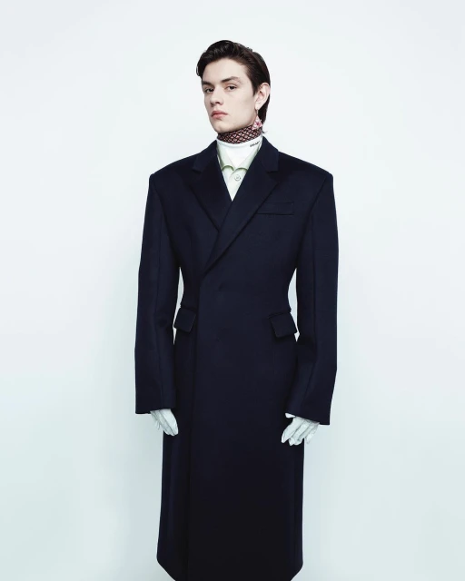 Fashion portrait of a young adult male model in an oversized navy coat, white gloves, and patterned neck scarf against a white backdrop.