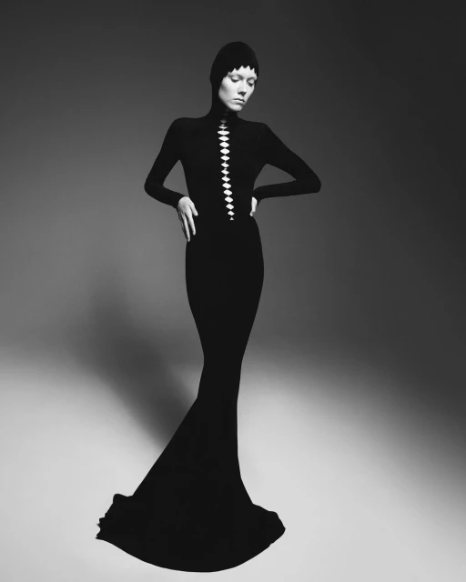 A fashion shot of a slender woman in a full-length black dress and hood, posing with hands on hips in a grey studio setting.
