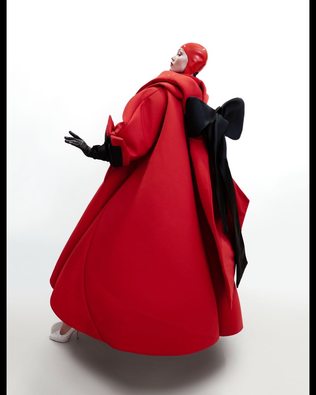 Elegant woman in a red couture dress with a large black bow, high heels and gloves, posing against a bright white background in a fashion shot.