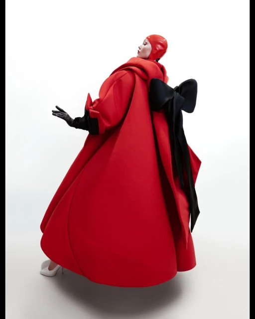 Elegant woman in a red couture dress with a large black bow, high heels and gloves, posing against a bright white background in a fashion shot.