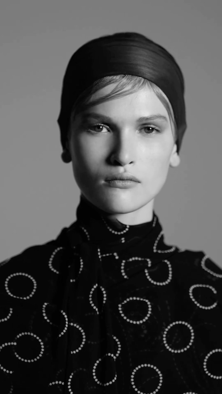 Black and white portrait of a woman with a dark headscarf and a dotted high-necked blouse looking directly at the camera in a studio.