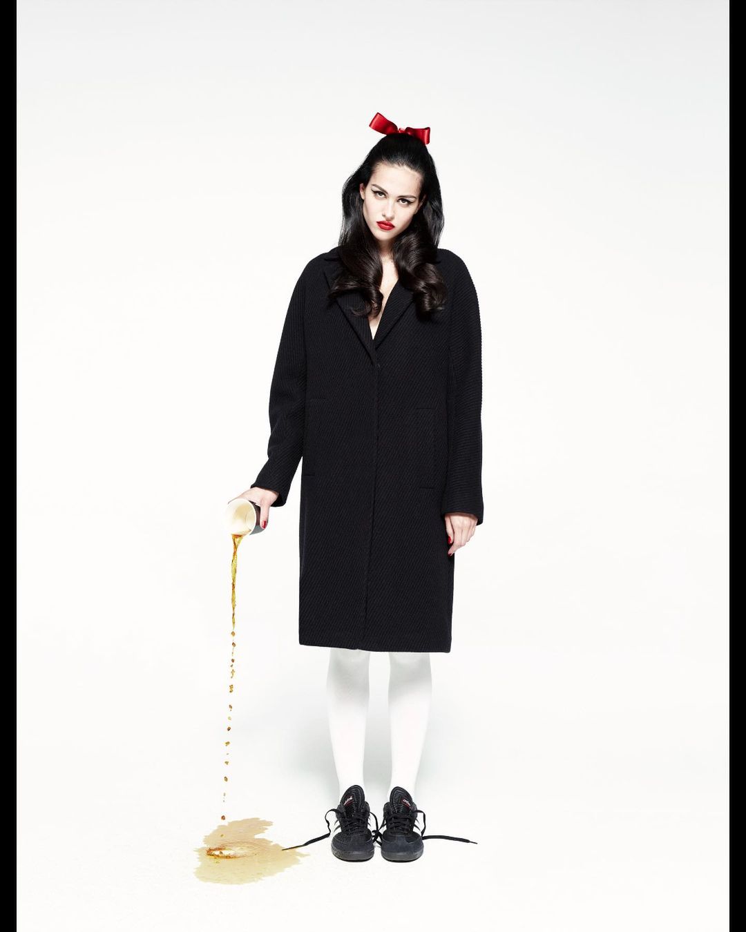A stylish young woman with a bow poses in front of a white backdrop while intentionally spilling liquid from a can onto the floor.