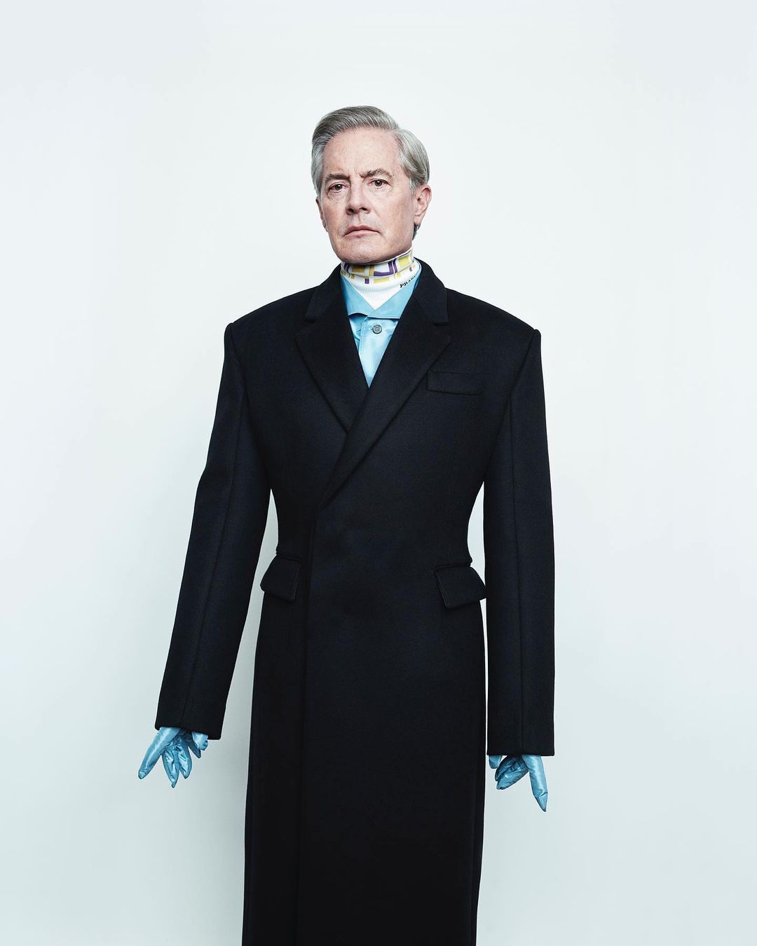 Portrait of adult male with grey hair wearing a dark-colored overcoat, light blue collared shirt, patterned neck scarf, and light blue gloves.