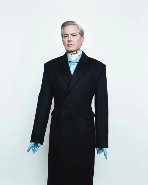 Portrait of adult male with grey hair wearing a dark-colored overcoat, light blue collared shirt, patterned neck scarf, and light blue gloves.