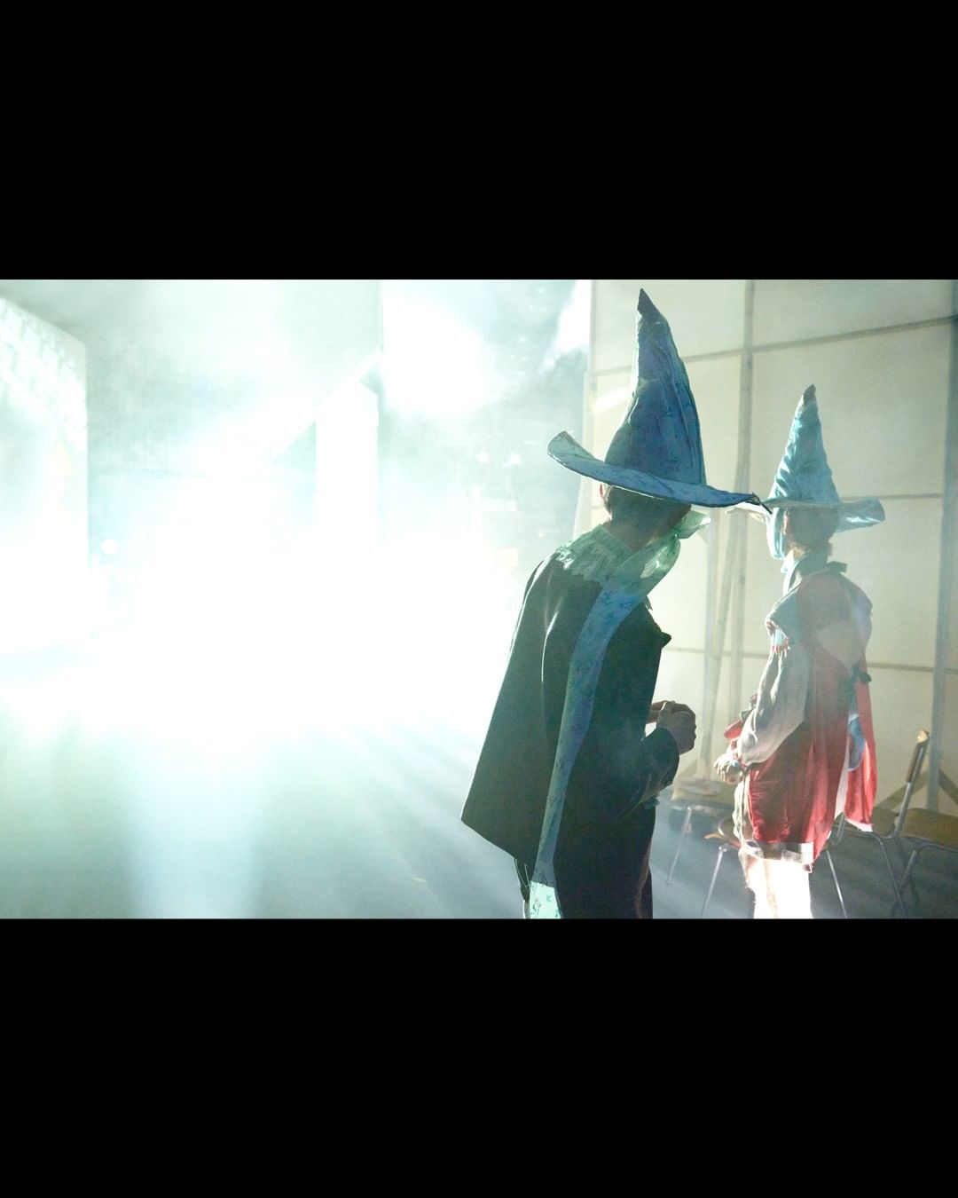 Two people in wizard costumes with conical hats and robes stand in a bright, hazy indoor space. One faces back, the other faces away.