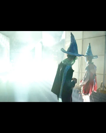 Two people in wizard costumes with conical hats and robes stand in a bright, hazy indoor space. One faces back, the other faces away.