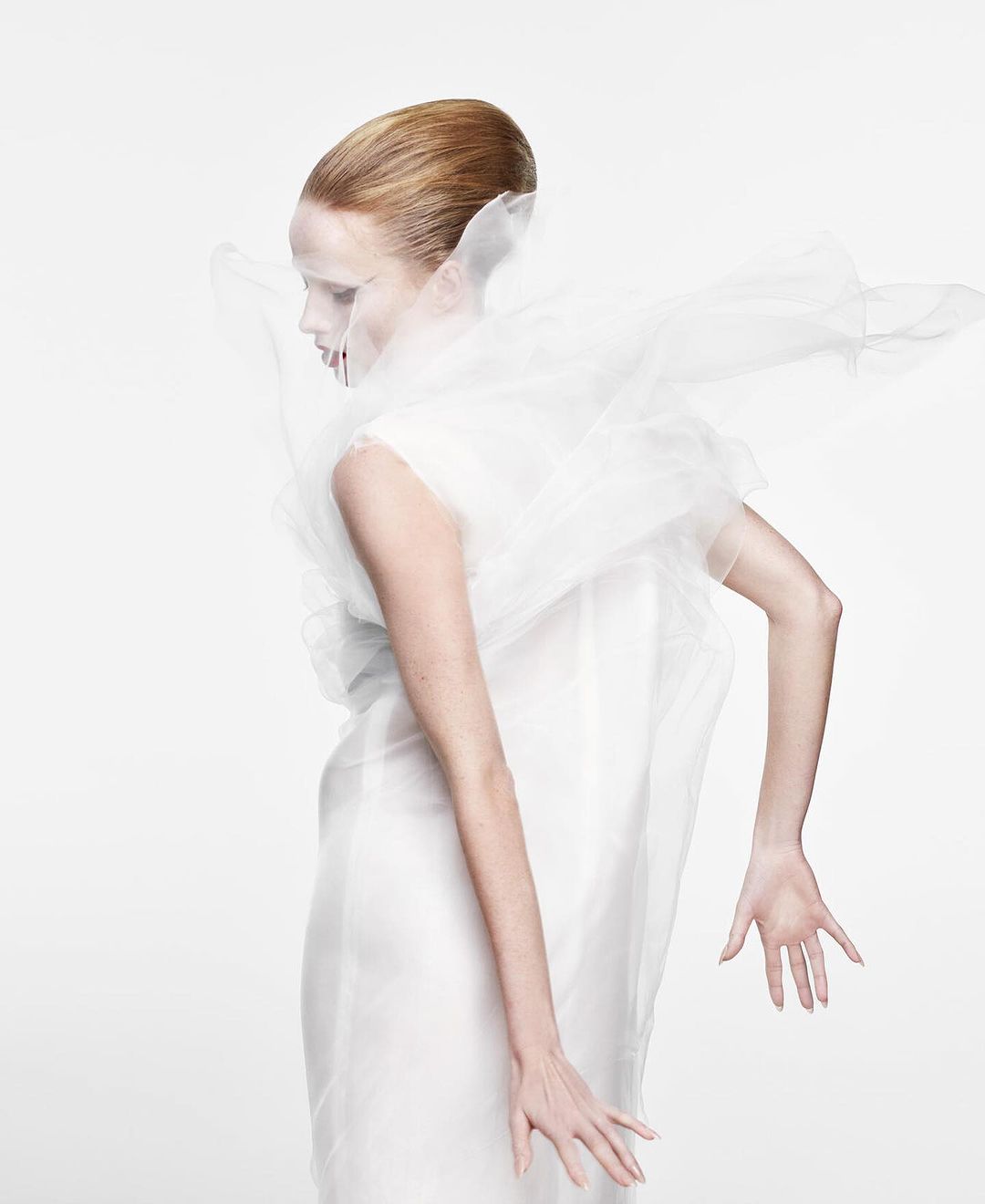 Elegant woman in white dress and translucent ruffled collar poses in a brightly lit studio, showcasing fashion and beauty.