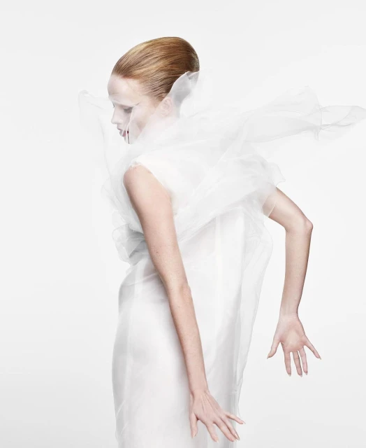 Elegant woman in white dress and translucent ruffled collar poses in a brightly lit studio, showcasing fashion and beauty.