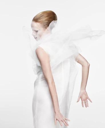 Elegant woman in white dress and translucent ruffled collar poses in a brightly lit studio, showcasing fashion and beauty.