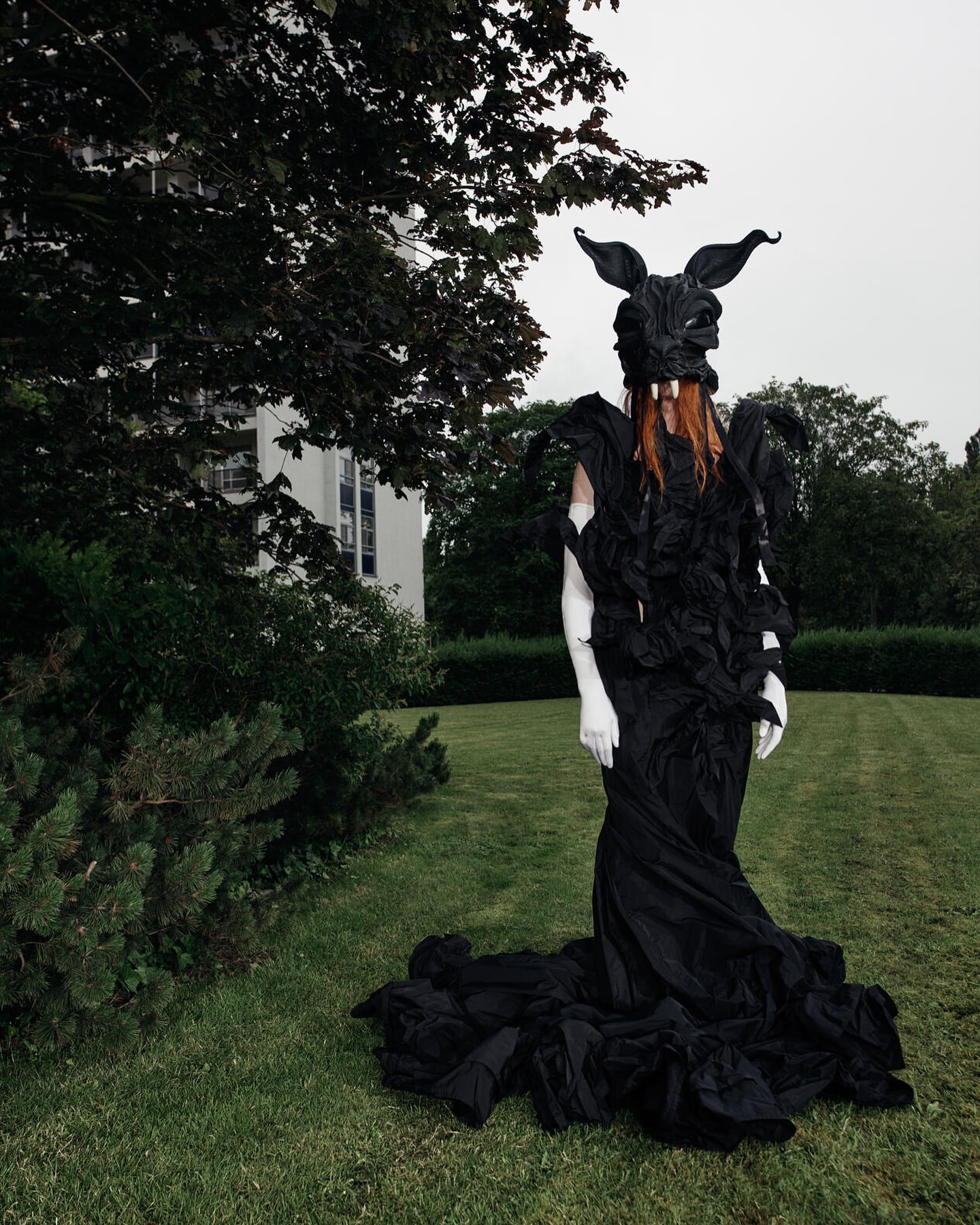 Model in elaborate black costume with horned rabbit mask standing on green grass before a white building.