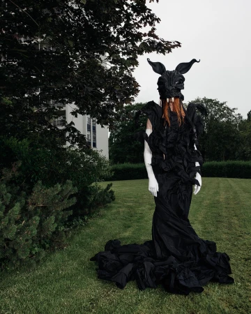 Model in elaborate black costume with horned rabbit mask standing on green grass before a white building.