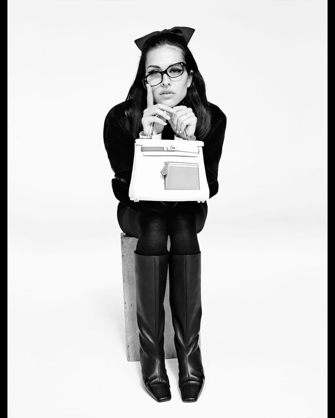 Black and white portrait of a woman with glasses and a hair bow sitting on a stool holding a handbag in a photo studio setting.