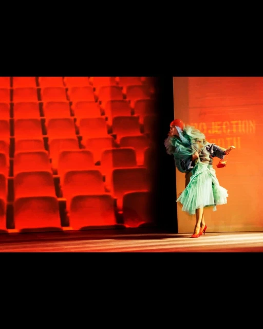 Person in teal dress and red heels stands on stage in front of red seating and stage lighting, creating a theatrical or performance setting.