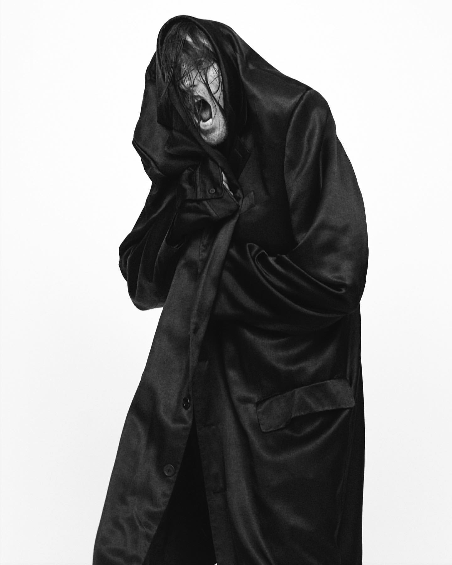 Monochrome portrait of a person with long hair wearing a hooded satin robe, looking down with an open mouth expression against white.