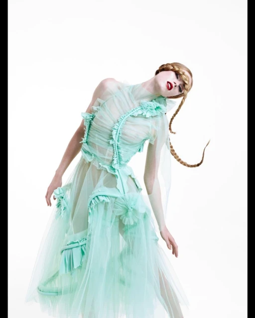 A woman poses wearing a sheer mint green tulle dress with blonde braids against a white background in a fashion editorial-style shot.