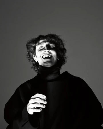 Monochrome portrait of a smiling person wearing a black turtleneck against a dark grey seamless studio background.