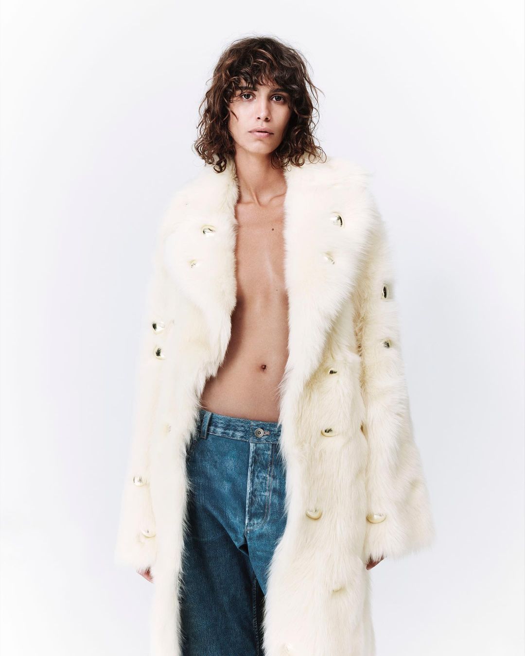 Person with brown curly hair poses in a long, creamy white fur coat with gold embellishments and blue denim jeans against a white background.