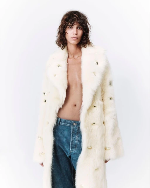 Person with brown curly hair poses in a long, creamy white fur coat with gold embellishments and blue denim jeans against a white background.