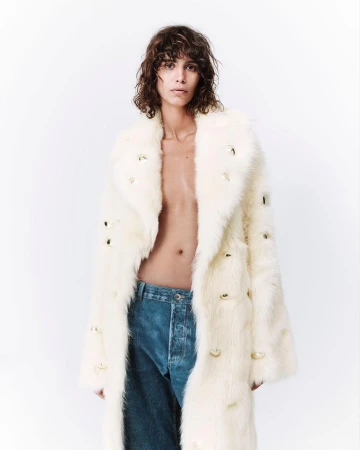 Person with brown curly hair poses in a long, creamy white fur coat with gold embellishments and blue denim jeans against a white background.