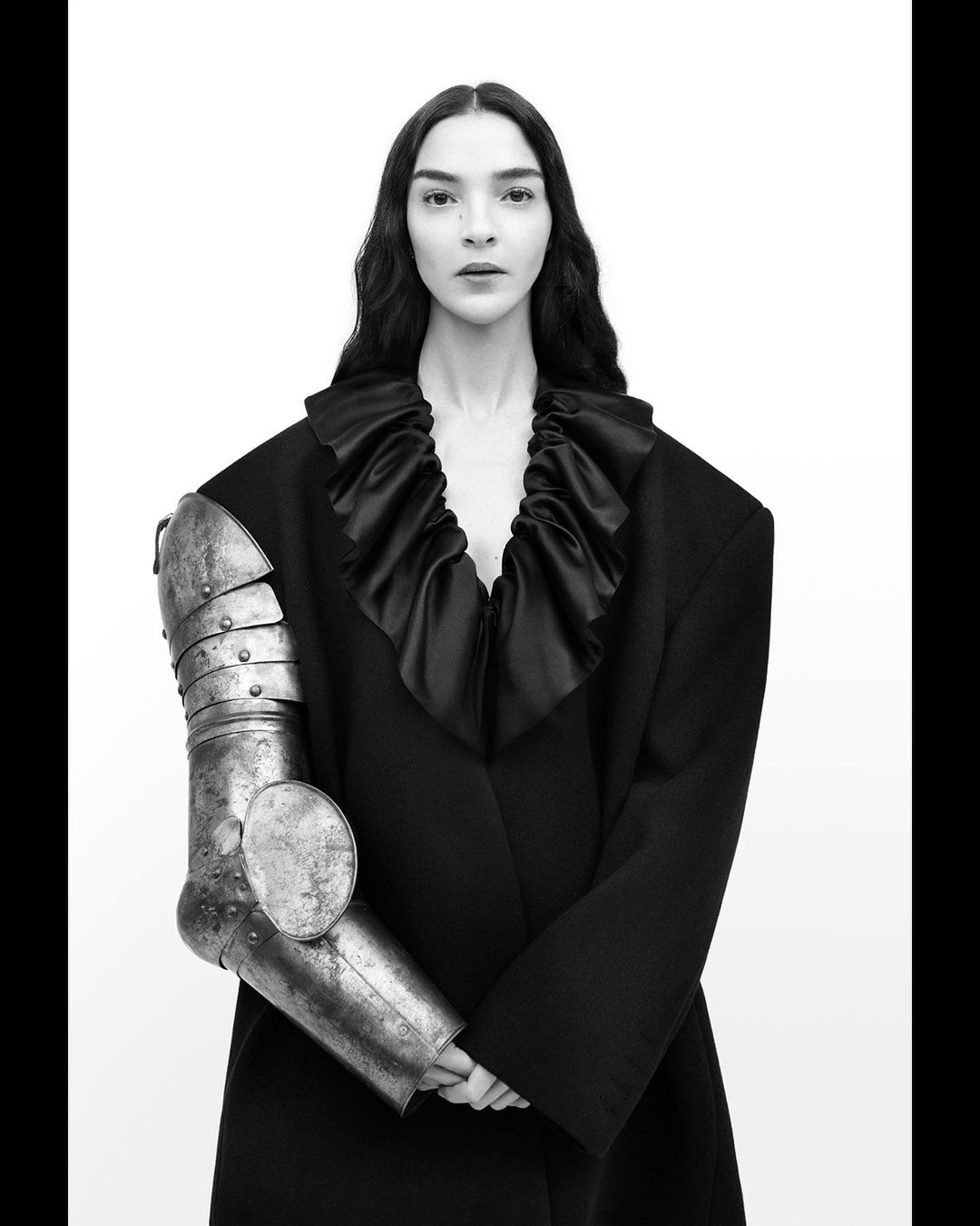 Black and white portrait of a woman in a black dress, wearing a single metal armor sleeve, against a bright white background.