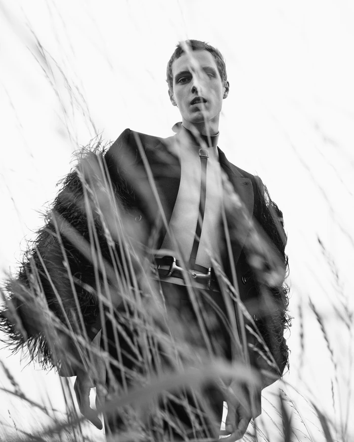 Monochrome portrait of a male model framed by tall grass wearing a structured jacket and harness by Willy Vanderperre.