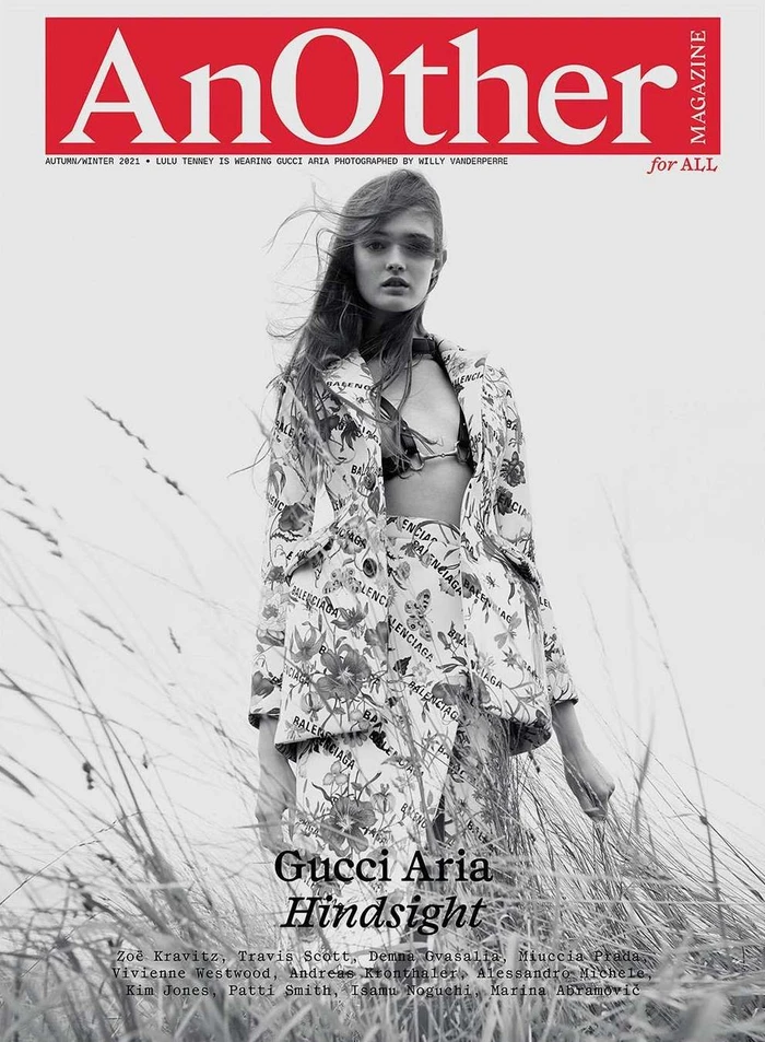 Model standing outdoors among tall grass wearing a patterned floral jacket and matching skirt photographed in black and white.