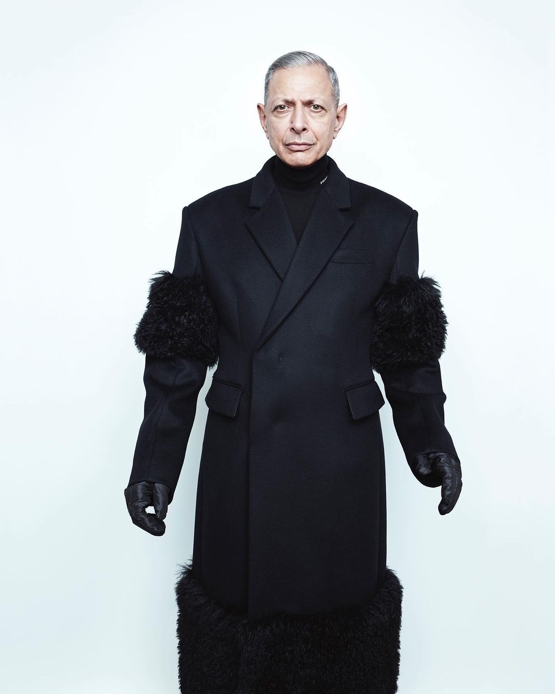 Jeff Goldblum wearing a long black coat with fur trim and black gloves against a plain white background