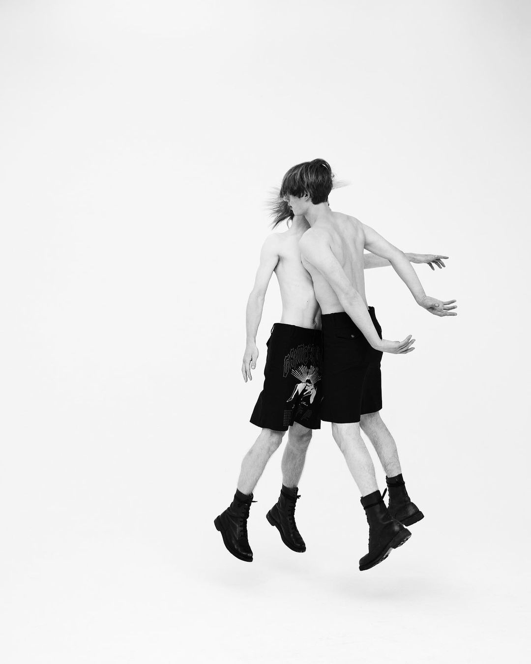 Two shirtless male models mid-air wearing black shorts and boots against a seamless white background.