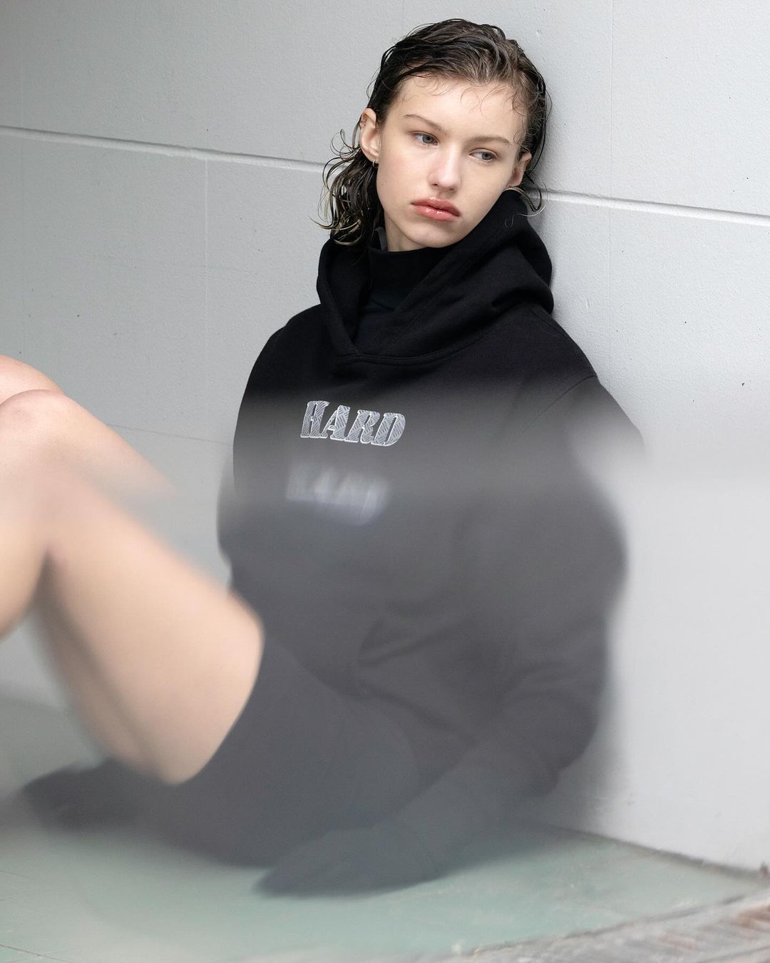 Indoor portrait of a young, light-skinned woman in black hooded sweatshirt and cycling shorts, sitting against a pale wall in studio.