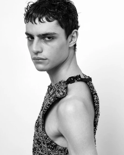 Black and white close-up studio portrait of a young adult male fashion model with dark curly hair wearing a tweed sleeveless top.