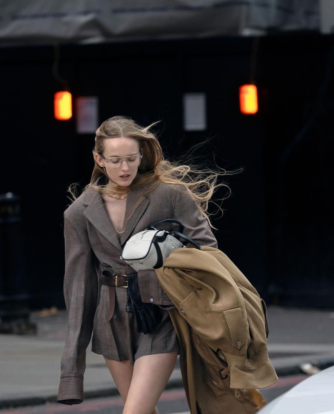 A fashionable adult female walks outdoors wearing a brown blazer and shorts outfit, glasses, and carrying a jacket and purse.