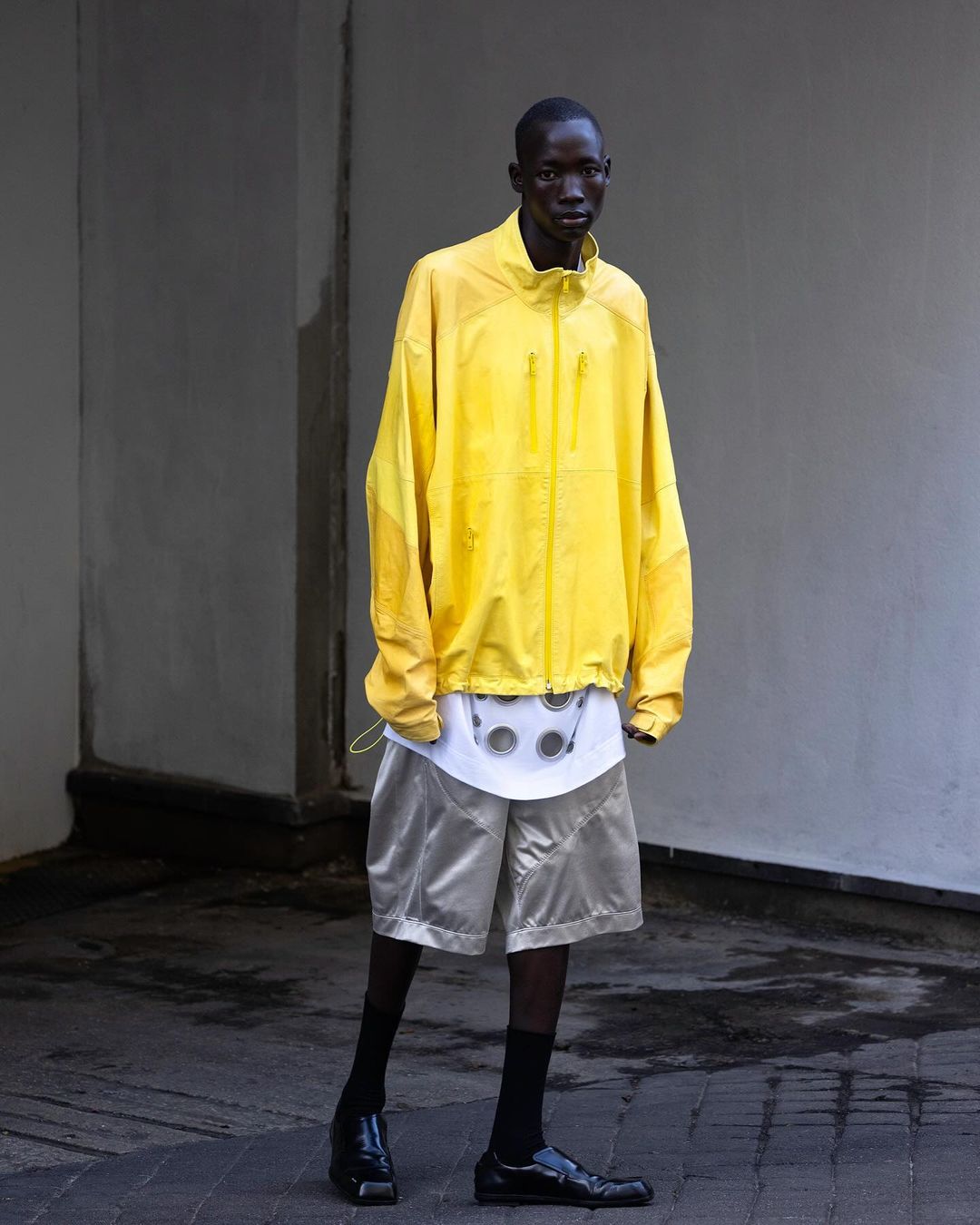 Man in a yellow jacket and shorts standing in front of a gray wall, looking at the camera. The image features a fashion-forward style.