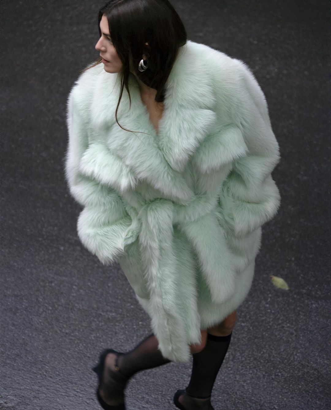 Stylish adult woman in a light green fur coat walking in an urban setting; fashion-forward attire and confident demeanor on a city street.