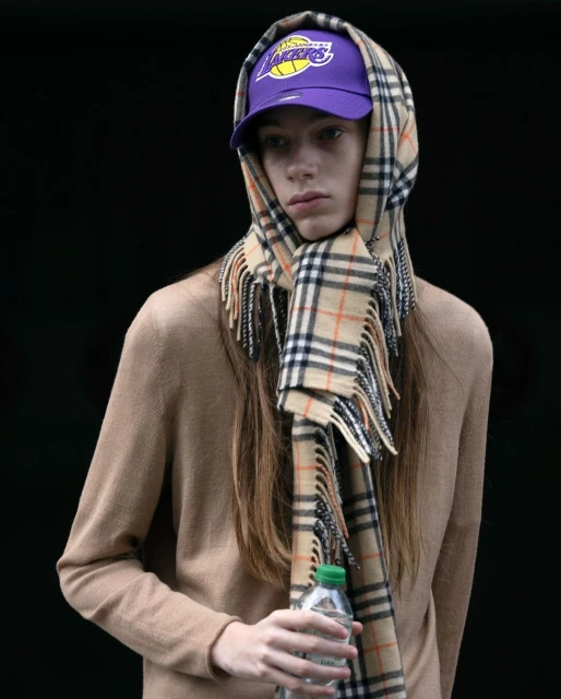 Adult wears a beige sweater, purple Lakers cap, and Burberry scarf while holding a plastic bottle against a black background in a portrait shot.