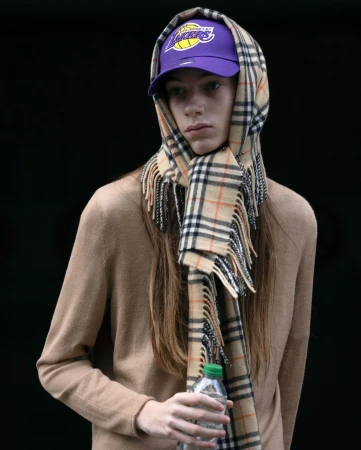 Adult wears a beige sweater, purple Lakers cap, and Burberry scarf while holding a plastic bottle against a black background in a portrait shot.