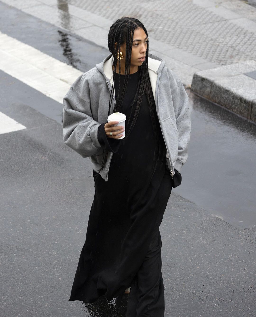 Adult female with braids in a gray hooded jacket and long black dress walks on a wet city sidewalk holding a white paper cup.