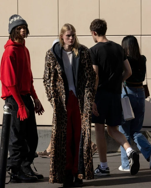 Fashionable people stand on a sidewalk wearing a red hoodie and long leopard print coat in an outdoor, sunny urban setting.