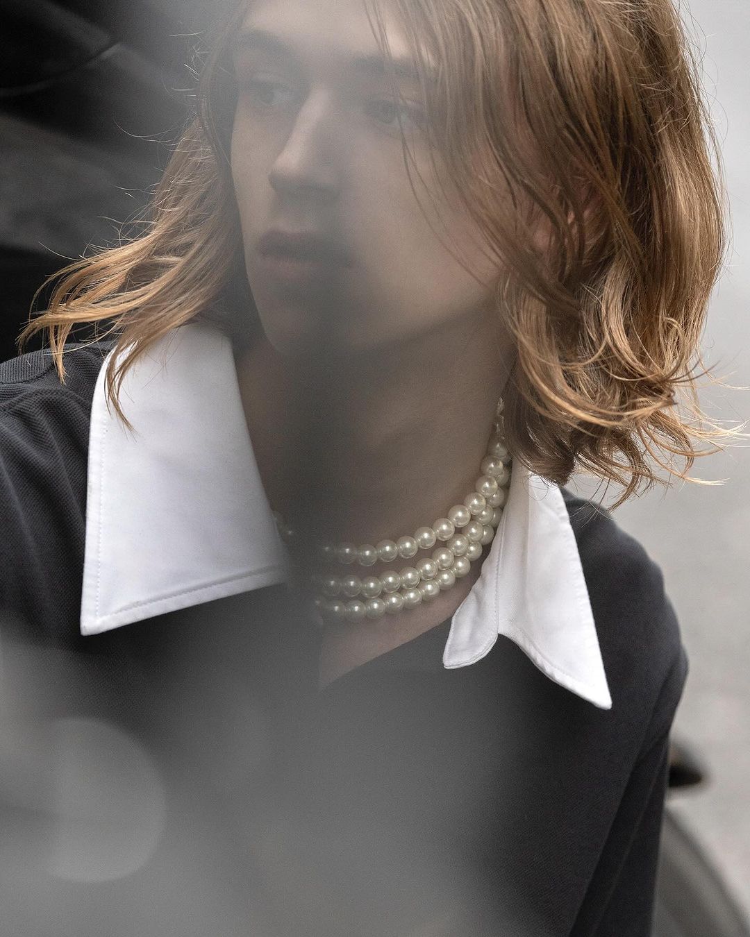 Close-up portrait of a young man with long auburn hair wearing a white collared shirt and layered pearl necklace by Winter Vandenbrink.