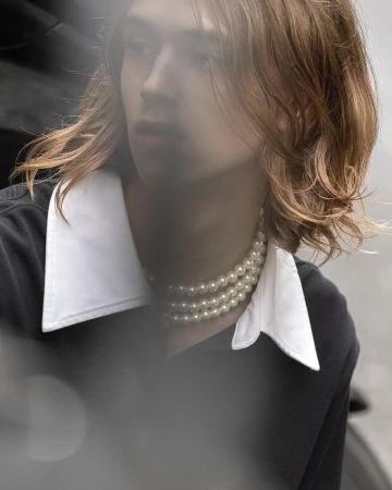 Close-up portrait of a young man with long auburn hair wearing a white collared shirt and layered pearl necklace by Winter Vandenbrink.