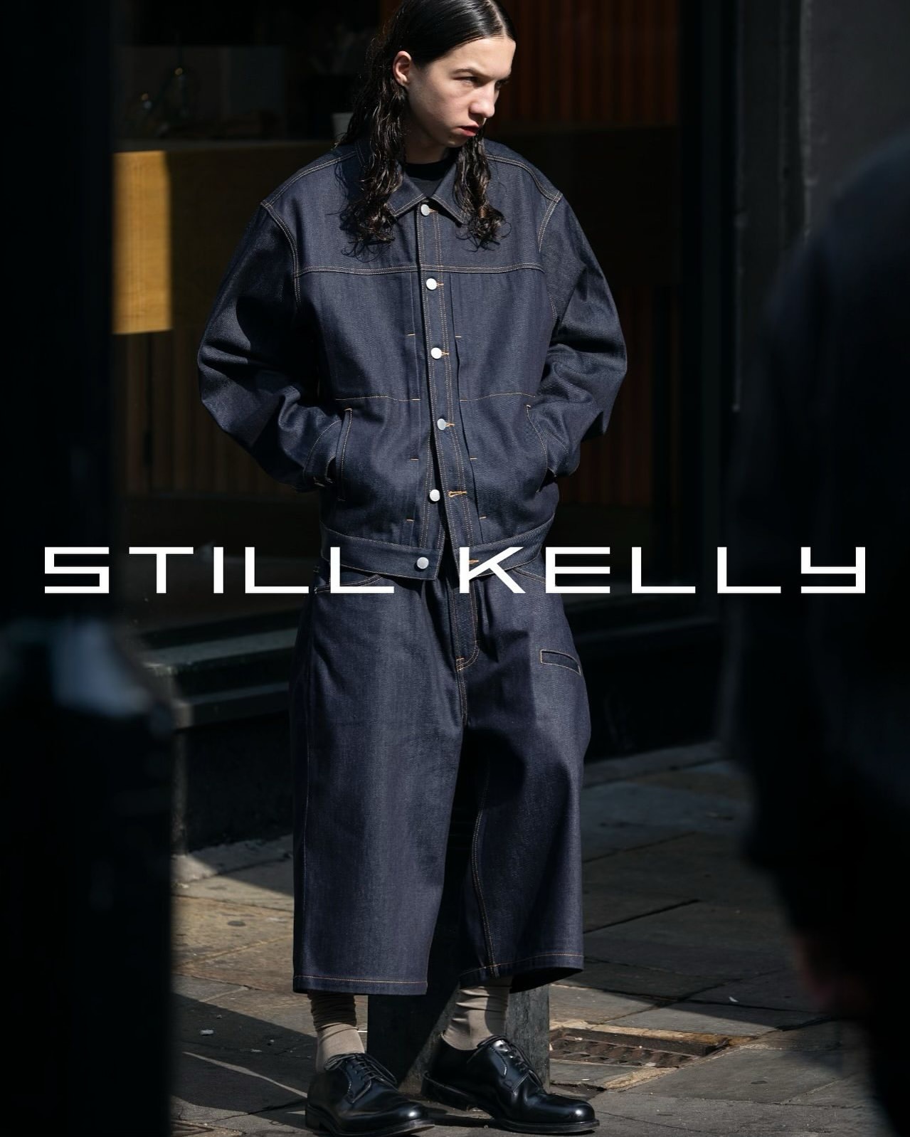 A young adult in a stylish denim jacket and wide-leg jeans stands outdoors, accented by 'STILL KELLY' text overlay, in a fashion-forward shot.