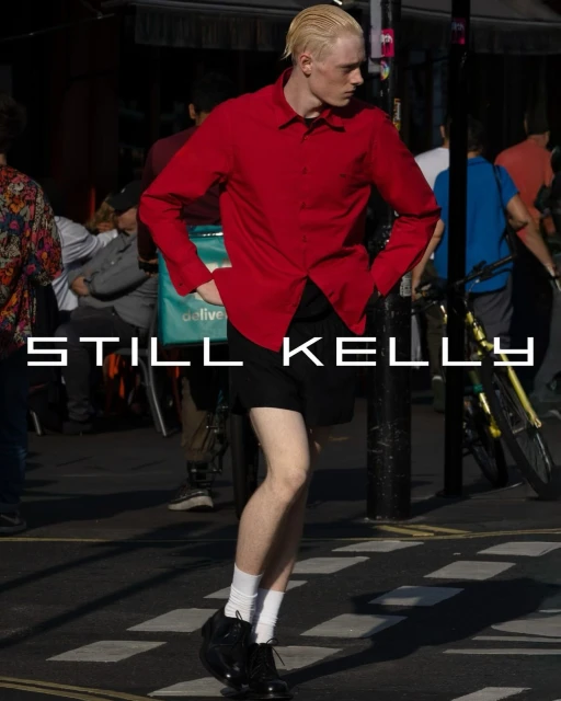 Fashionable adult male walks on a city street wearing a red button-down shirt, black shorts, white socks and black dress shoes in daylight.