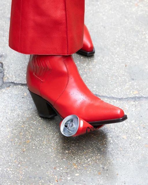 Close-up of red cowboy boots standing next to a crushed red and silver aluminum can on concrete. The boots match the pants.
