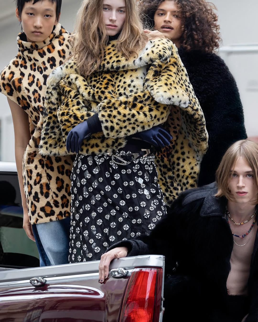 Five models showcase fashion designs including faux fur coats and patterned skirt, leaning against a truck with an urban background. 