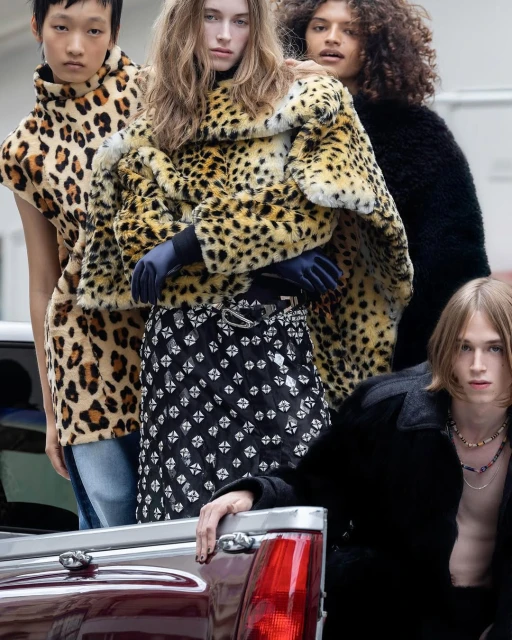Five models showcase fashion designs including faux fur coats and patterned skirt, leaning against a truck with an urban background. 