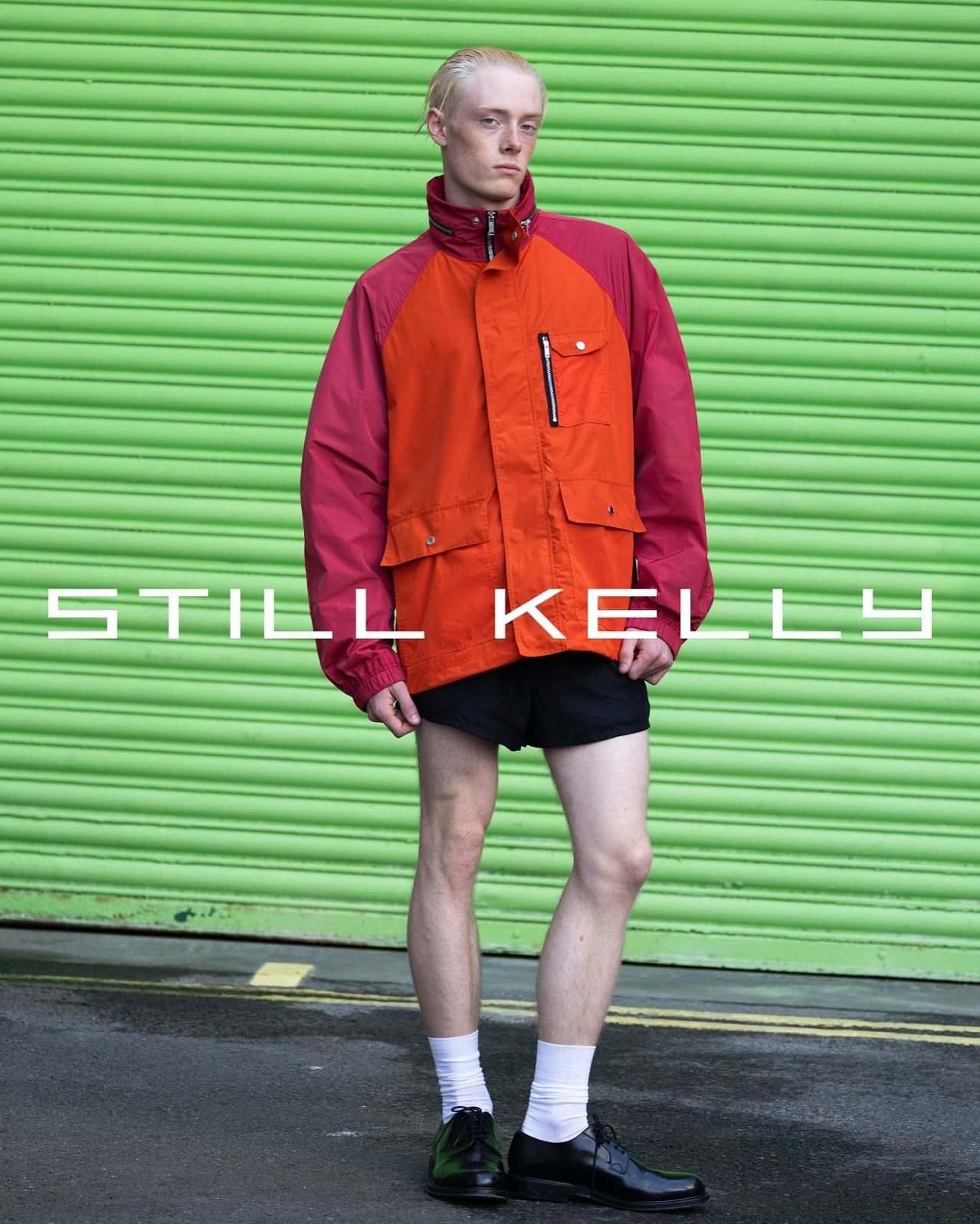 Full body shot of a slender male with blond hair in an orange and magenta jacket, black shorts, white socks, and black dress shoes.