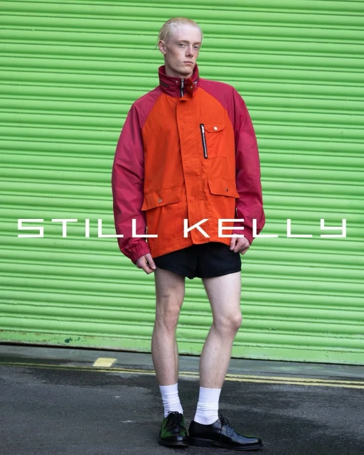 Full body shot of a slender male with blond hair in an orange and magenta jacket, black shorts, white socks, and black dress shoes.