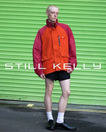 Full body shot of a slender male with blond hair in an orange and magenta jacket, black shorts, white socks, and black dress shoes.