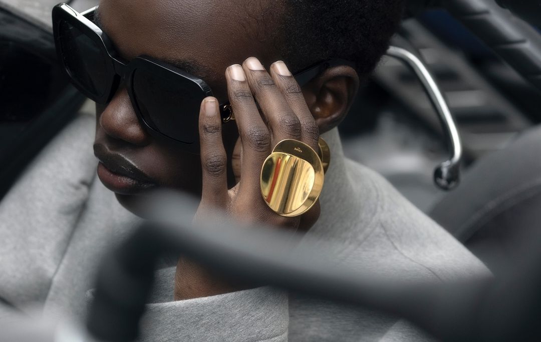 Close-up of a black adult woman wearing sunglasses and gold rings on her hand, with a grey sweater, and dark background.